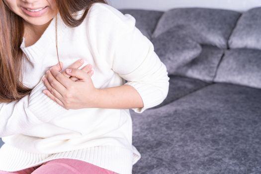 Young woman suffering from heart pain while sitting on sofa at home. Healthcare medical or daily life concept.