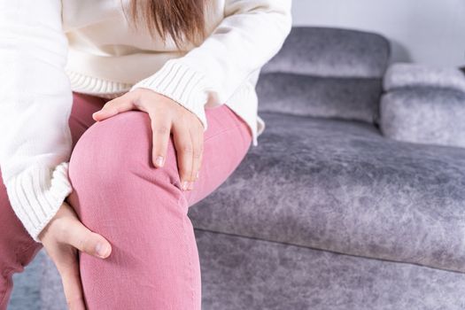 Young woman suffering from knee pain while sitting on sofa at home. Healthcare medical or daily life concept.