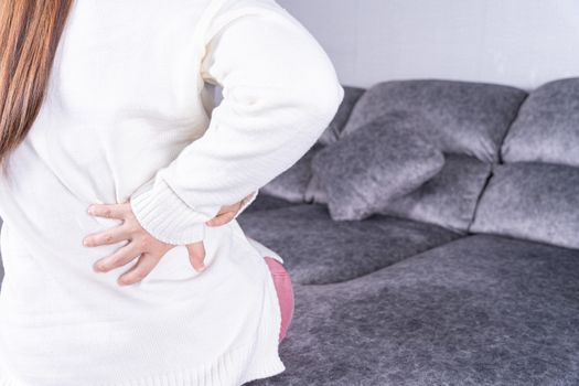 Young woman suffering from back pain while sitting on sofa at home. Healthcare medical or daily life concept.