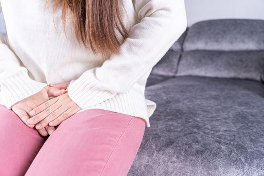 Woman hands holding her crotch suffering from painful stomachache while sitting on sofa at home. Healthcare medical or daily life concept.