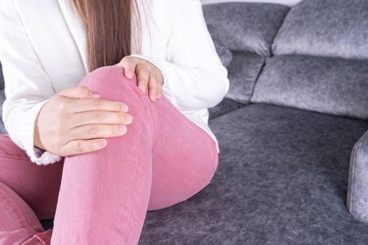 Young woman suffering from knee pain while sitting on sofa at home. Healthcare medical or daily life concept.