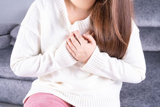 Young woman suffering from heart pain while sitting on sofa at home. Healthcare medical or daily life concept.