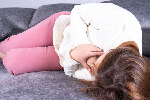 Young woman suffering from heart pain while sitting on sofa at home. Healthcare medical or daily life concept.