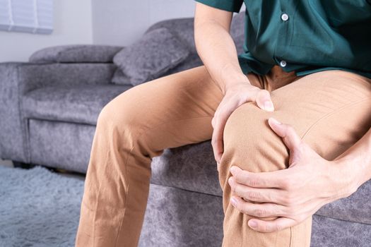 Young man suffering from knee pain while sitting on sofa at home. Healthcare medical or daily life concept.