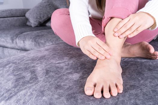Young woman suffering from ankle pain while sitting on sofa at home. Healthcare medical or daily life concept.