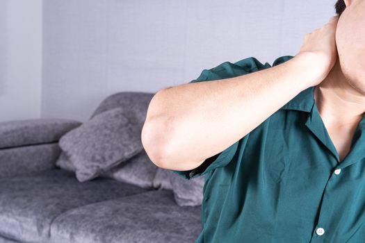 Young man suffering from neck pain while sitting on sofa at home. Healthcare medical or daily life concept.