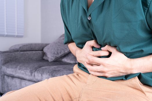 Young man suffering from abdominal pain while sitting on sofa at home. Healthcare medical or daily life concept.