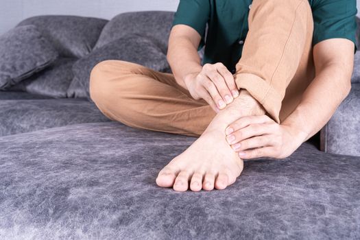 Young man suffering from ankle pain while sitting on sofa at home. Healthcare medical or daily life concept.
