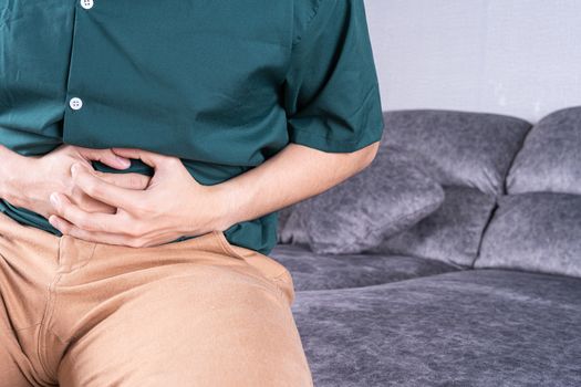 Young man suffering from abdominal pain while sitting on sofa at home. Healthcare medical or daily life concept.