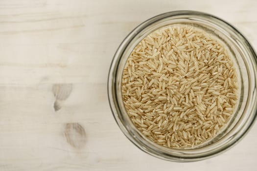 Back to the natural vegan food theme copy space: top down flat lay view of a big glass jar filled of raw thaibonnet brown rice grains on light wooden table background