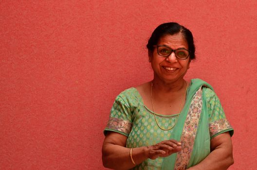 Happy looking senior Indian lady wearing traditional suit and spectacles standing against a pink wall in pink city Jaipur, Rajasthan, India