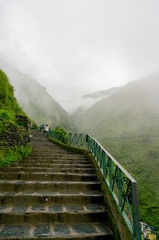 a series of stairs leading to the bhagsu falls in McLeodganj in himachal india. The lush green plants, foggy misty clouds in distance shows the famous triund trek of Himachal pradesh in India