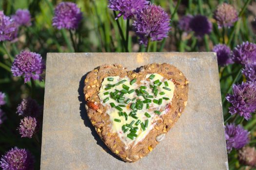 The picture shows a heartshaped bread slice in front of blossoming chives