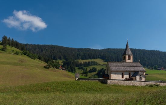 South Tyrolean mountain church under a blue sky with a single white cloud, mountain landscape with meadows and pines