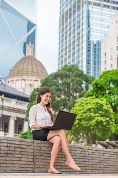 Asian woman using computer laptop wifi online outside in city park working typing in Hong Kong. Young autonomous worker or student sitting outdoor, freelance work or digital nomad on Asia travel.