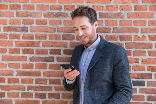 Young business man using smartphone in urban city background relaxing on brick wall texting sms on phone app living a modern lifestyle. Happy businessman playing mobile games.