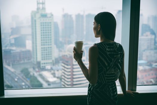Businesswoman drinking coffee at work contemplative looking out the window of high rise skyscraper building during morning tea break. Stress, mental health in the workplace. Career job concept.