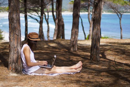 Freelance woman working in vacation on the laptop sitting in a pine forest on the sea, smart working lifestyle concept theme and life choice