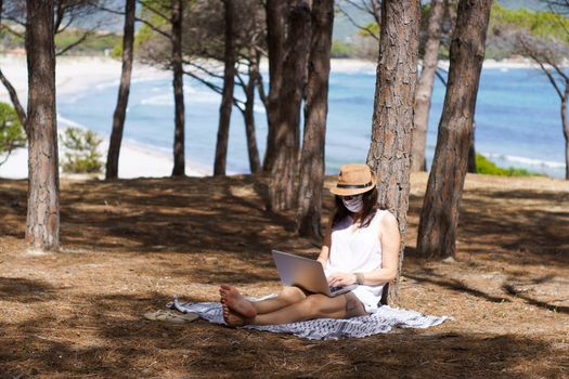 Freelance woman working in vacation on the laptop sitting in a pine forest on the sea with face mask protection for coronavirus, smart working lifestyle concept theme and life choice