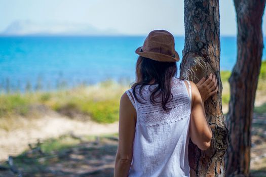 Pensive young woman with long hair and hat, seen from behind, looks at the sea leaning against a tree in a pine forest