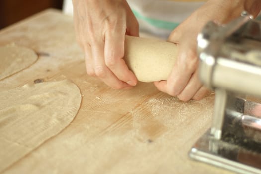 Making homemade fresh pasta: angle view of woman's hands kneading fresh pasta dough on the wooden work table with flour, dough, and metal manual fresh pasta machine