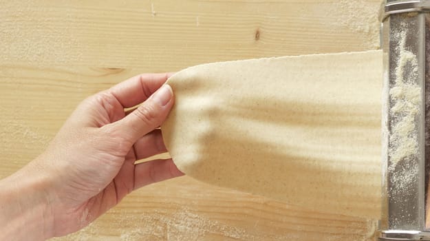 Making fresh homemade pasta: overhead top view of woman's hand pulling out the rolled dough from the manual pasta making machine backlit