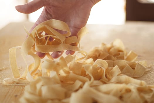Preparing fresh homemade pasta: close-up view of woman's hand pulling up some fresh tagliatelle on wooden work table in natural sunlight