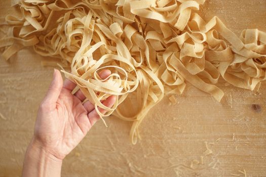 Preparing fresh homemade pasta: overhead close-up view of woman's hand pulling up some fresh tagliatelle on wooden work table in natural sunlight