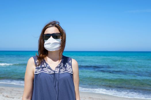 Coronavirus seaside holidays: a woman standing on the sand at the beach looking at the sun with the mask for Covid-19 pandemic