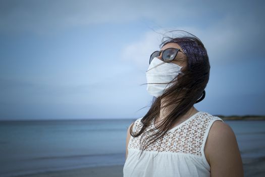 Coronavirus seaside holidays: half-length shot of a woman at the beach looking at the sun with the mask for Covid-19 pandemic