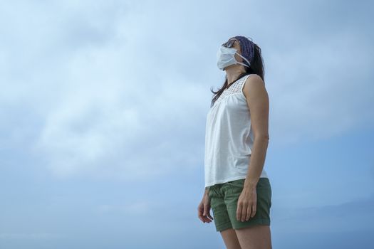 Coronavirus seaside holidays: shot of a woman at the beach looking at the sun with the mask for Covid-19 pandemic with cloudy sky