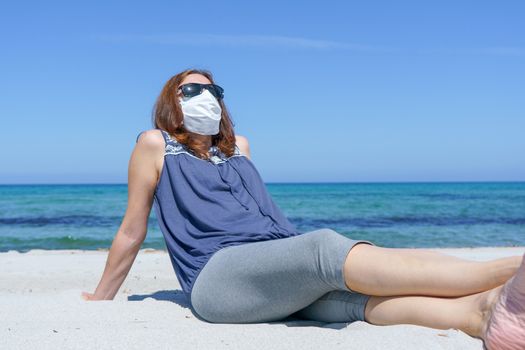 Coronavirus seaside holidays: a woman sitting on the sand at the beach look at the sun with the mask for Covid-19 pandemic