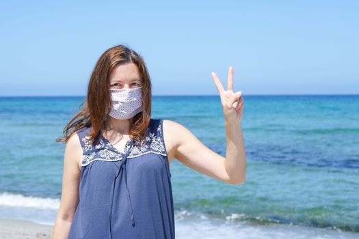 Coronavirus seaside holidays: a woman standing on the sand at the beach looking at the camera with fingers in victory and the mask for Covid-19 pandemic