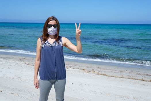 Coronavirus seaside holidays: a woman standing on the sand at the beach looking at the camera with fingers in victory and the mask for Covid-19 pandemic