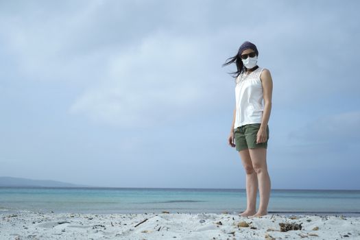 Coronavirus seaside holidays: shot of a woman at the beach look at the camera with the mask for Covid-19 pandemic with cloudy sky