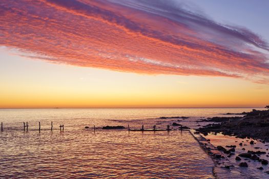 Orange and red sky with gorgeous cloud on an old abandoned sea pier in dawn or sunset with water reflections between the rocks