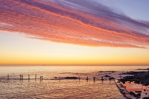 Orange and red sky with gorgeous cloud on an old abandoned sea pier in dawn or sunset with water reflections between the rocks