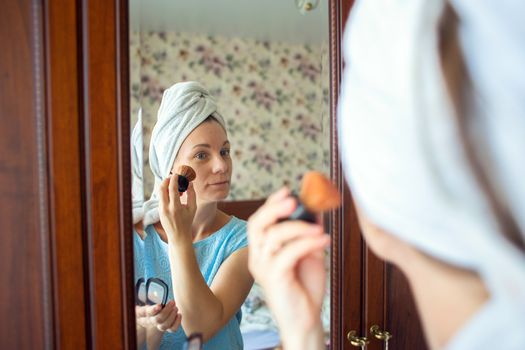 A woman with a washed head in a towel does makeup in her bedroom in the morning. Girl puts a blush on her cheeks with a brush