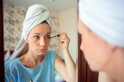 A woman with a washed head in a towel does makeup in her bedroom in the morning. Girl paints eyelashes with mascara