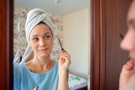 A woman with a washed head in a towel does makeup in her bedroom in the morning. Girl paints eyelashes with mascara