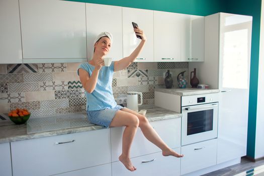 A woman with a towel on her head after a shower sits in her kitchen with a cup of coffee or tea and photographs herself on a smartphone.