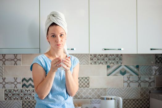 A woman washed her hair in the morning and drinks coffee with a towel on her head. Morning toilet and breakfast in the kitchen