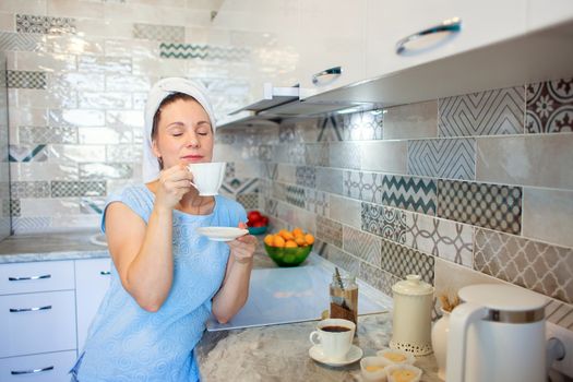 A woman washed her hair in the morning and drinks coffee with a towel on her head. Morning toilet and breakfast in the kitchen