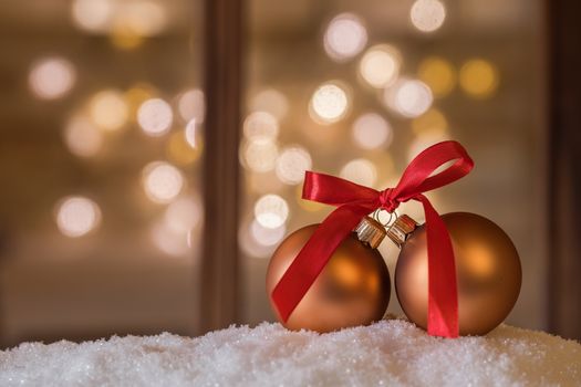 Christmas baubles with red bow on snow in front of window with blurred sparkling lights background