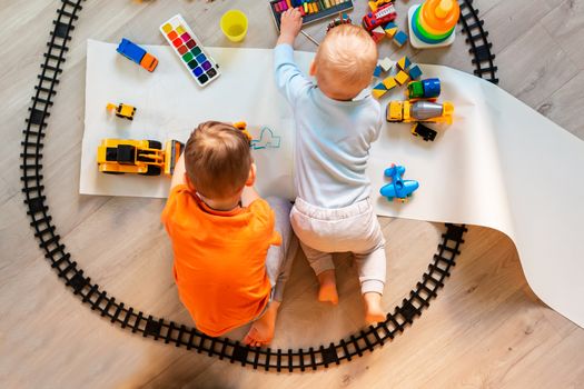 Preschool boys drawing on floor on paper, playing with educational toys - blocks, train, railroad, vehicles at home or daycare. Toys for preschool and kindergarten. Top view. Children's art and creativeness concept