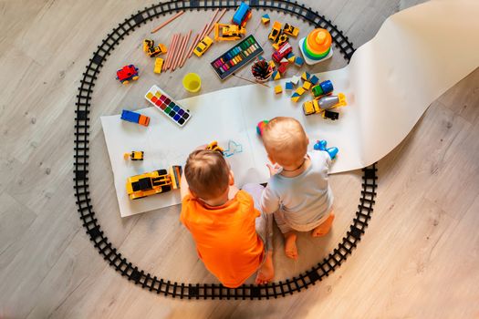 Preschool boys drawing on floor on paper, playing with educational toys - blocks, train, railroad, vehicles at home or daycare. Toys for preschool and kindergarten. Top view. Children's art and creativeness concept