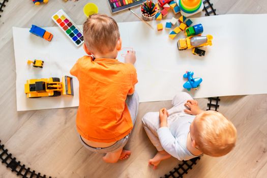 Preschool boys drawing on floor on paper, playing with educational toys - blocks, train, railroad, vehicles at home or daycare. Toys for preschool and kindergarten. Top view. Children's art and creativeness concept