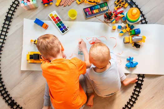 Preschool boys drawing on floor on paper, playing with educational toys - blocks, train, railroad, vehicles at home or daycare. Toys for preschool and kindergarten. Top view. Children's art and creativeness concept