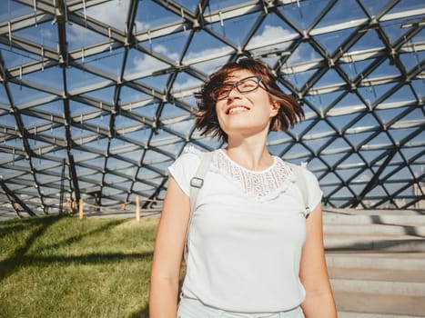 Wide smiling woman shakes her hair. Woman under modern dome made of transparent glass. City life.
