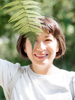 Young woman is hiding her eyes with fern leaf. Symbol of life, tranquility and unity with nature. Summer in forest.